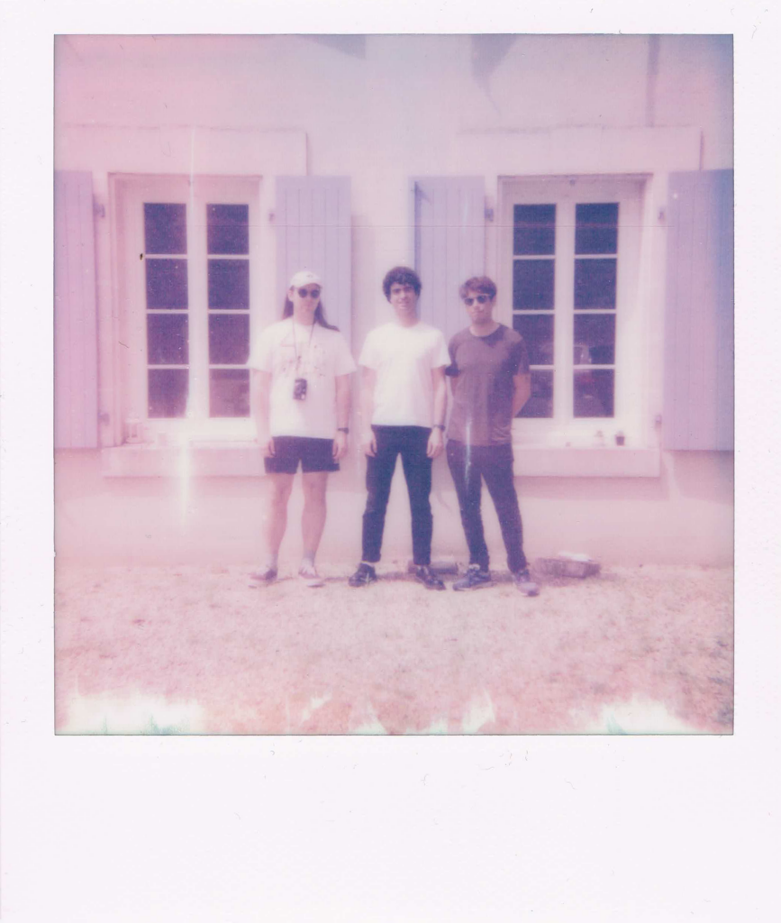 A bleached out Polaroid of the band Adventure Team's three members standing in front of the old farm house in France where they recorded their 2nd album